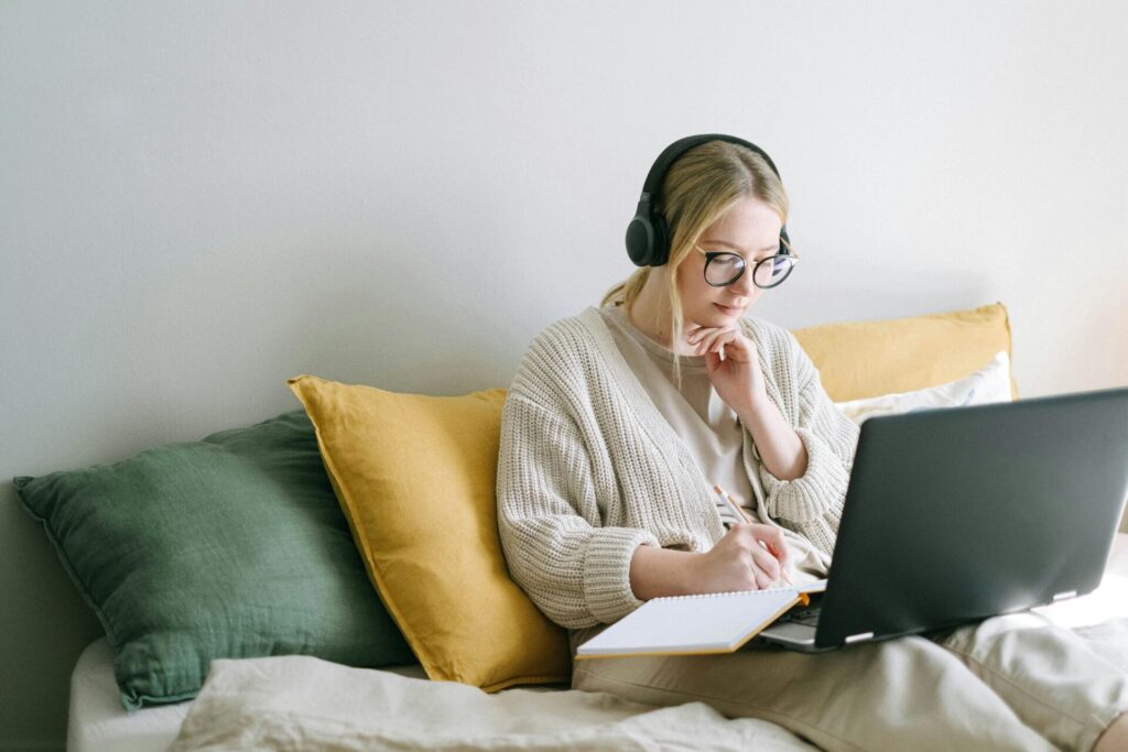 Une femme concentrée, portant des lunettes et un casque, travaille sur un ordinateur portable depuis un lit confortable.