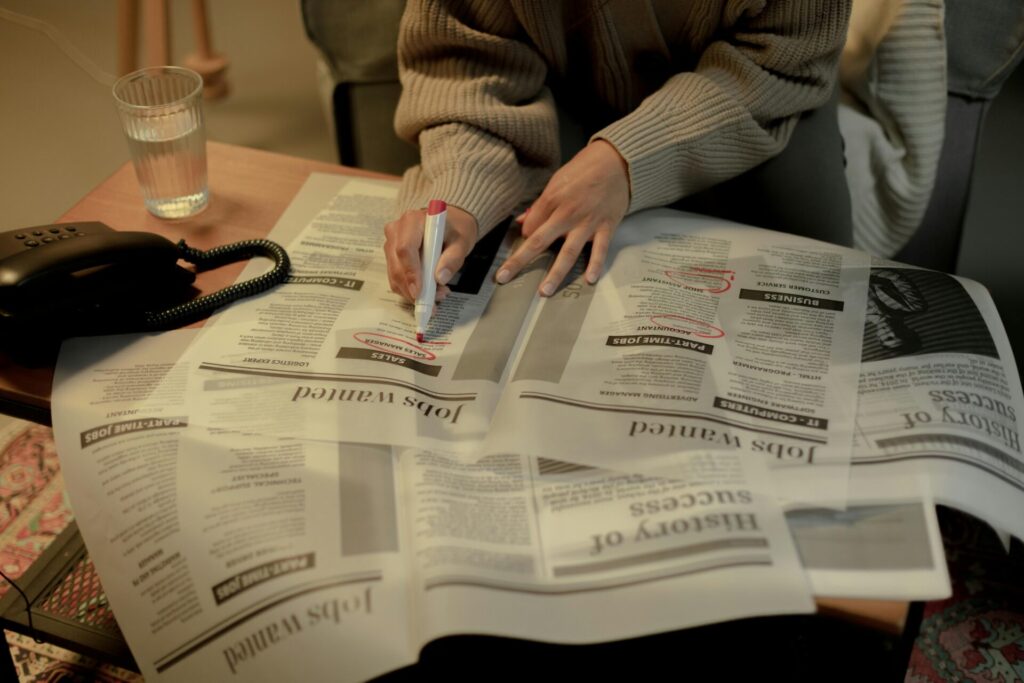 An adult woman marks job listings in a newspaper while sitting indoors, highlighting job search activity.